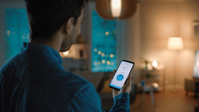 Young Handsome Man Gives a Voice Command to a Smart Home Application on His Smartphone To Switch on the Lights and Set a Comfortable Temperature. It's a Cozy Evening in Living Room.
